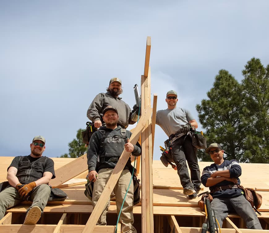 TM Construction carpenters taking a break on a roof they are framing, tool belts on, pines behind them